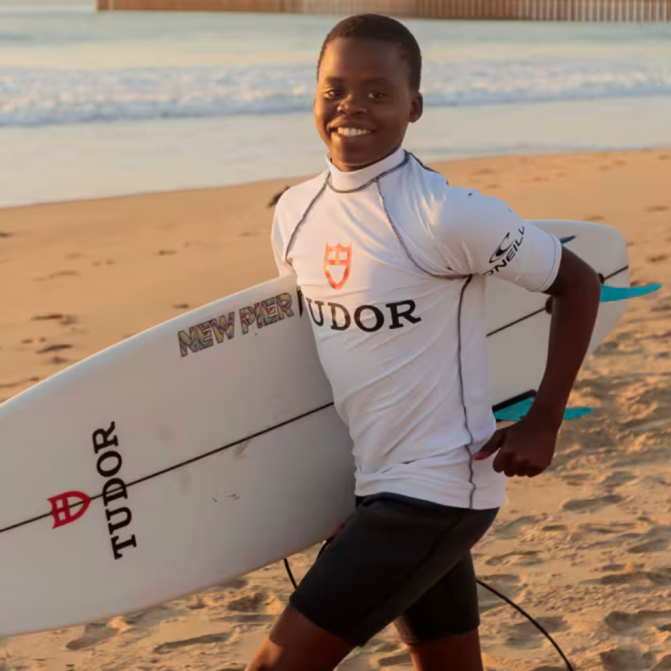 A smiling young boy stands on a sandy beach holding a white surfboard branded TUDOR and NEW PIER. He is wearing a white TUDOR surf rash guard and black swim trunks, with the ocean and sunset in the background.